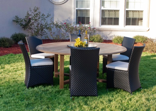 70256 Valencia 7 piece Dining Set of 4 wicker chairs and a 6 foot round teak dining table showing 2 wine bottles and yellow flowers on lazy susan in center on grass with house in back