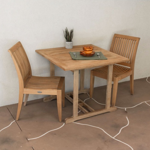 70303 Laguna 3 piece Square Dining Set of 2 teak side chairs and a square teak dining table with place setting and potted plant on table top on stone patio against a wall