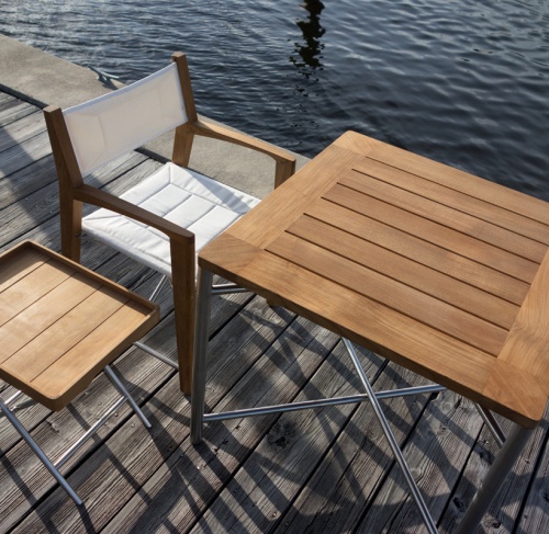 70460 Odyssey 3 piece Teak and Stainless Steel Set of Odyssey folding dining table and folding chair and folding ottoman with teak tray on wood deck and water in background
