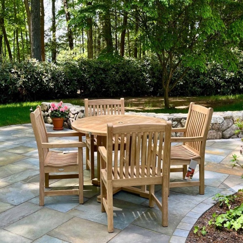 70483 Veranda teak 4 ft Round Dining Set of 4 Veranda Teak Armchairs and 4 foot round teak dining table on stone patio with plants and trees in background 