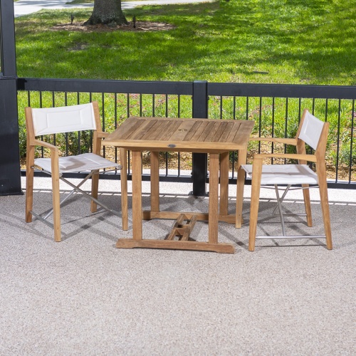 70651 Odyssey 3 piece Square Dinette Set of 2 folding director chairs and a teak 36 inch square dining table on patio against a black railing with grass and walkway and a tree in back