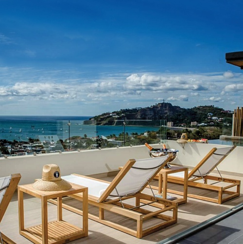 70780 Maya teak Sling Lounger Set on balcony with glass railing hat on side table silver ice bucket with champange on side table magazine on bottom shelf hat and fruit on ledge water view 