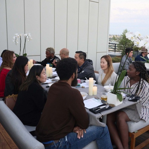 70883 Maya Horizon booth seat dining set with people sitting on set  pillar candles and potted white orchid plants on rooftop patio with bright sky in background