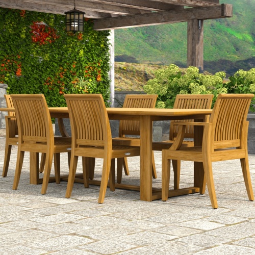70377 Horizon Laguna 7 pc Teak Dining Set of Horizon Dining Table and 2 Laguna DIning Arm Chairs and 4 Laguna Side Chairs on paver patio with raise flower bed with landscaping plants in the background