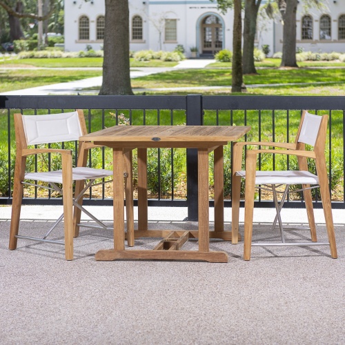 70651 Odyssey 3 piece Square Dinette Set of 2 folding director chairs and a teak 36 inch square dining table on patio with a black railing and grass and walkway and building in background