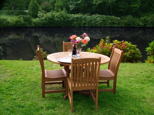 70054 Veranda Barbuda 5 piece Dining Set on green grass field with vase of flowers serving tray with wine bottle and 2 wine glasses on table overlooking lake and shrubs in background