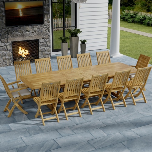 70059 Grand Barbuda 13 pc Teak Dining Set with optional seat cushions in angled aerial view on patio in front of an outdoor fireplace and potted plants with house and lawn in background