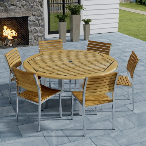 70439 Vogue Dining Set for 6 of Vogue Teak and Stainless Steel 60 inch Round Dining Table and 6 Vogue Side Dining Chairs angled top view with an outdoor fireplace in the background 