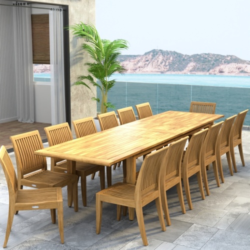 70917 Laguna 15 pc Teak Dining Set of Laguna 11 ft Rectangular Table and 14 Dining Side Chairs in angled end view on tiled patio with palm tree and land and ocean in background
