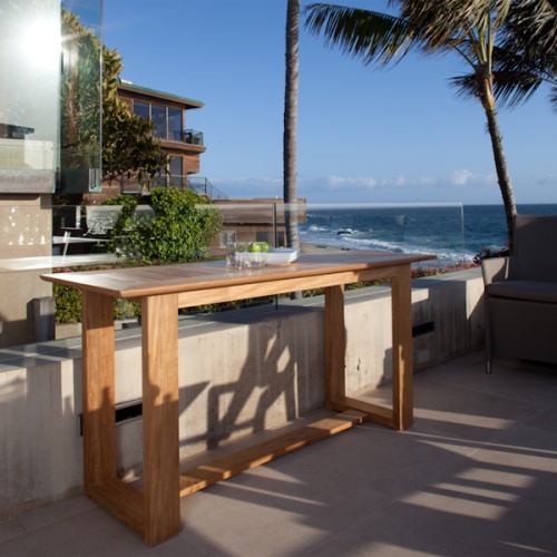 15669 Horizon Teak Console Table angled side view with 2 glasses and bowl of lime and 4 plates on outdoor terrace surrounded by palm trees and landscape plants overlooking ocean