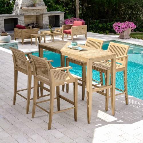 15821 Laguna 5 foot Teak Bar Table and 4 barstools on patio stones by pool with fireplace two potted flowering plants two teak Craftsman Chairs and shrubs in background