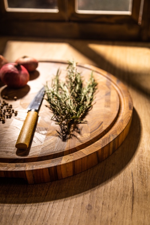 19122 Butcher Block 15 inch Round Charcuterie Cutting Board with rosemary and 3 red onions and whole black pepper and a knife with yellow handle partial aerial view on a counter top and window in background 