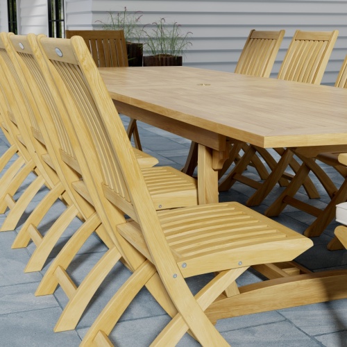 70059 Grand Barbuda 13 pc Teak Dining Set showing closeup end view of table and side chair on patio with plants in the background