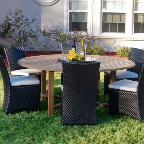 70256 Valencia 7 piece Dining Set of 4 wicker chairs and a 6 foot round teak dining table showing 2 wine bottles and yellow flowers on lazy susan in center on grass with house in back 