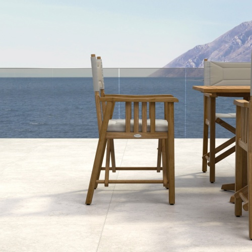 70360 Pyramid Director 7 piece teak Dining Set of 6 teak Director Chairs and Pyramid Teak Dining Table end view on stone patio in front of 2 palm trees and land in background