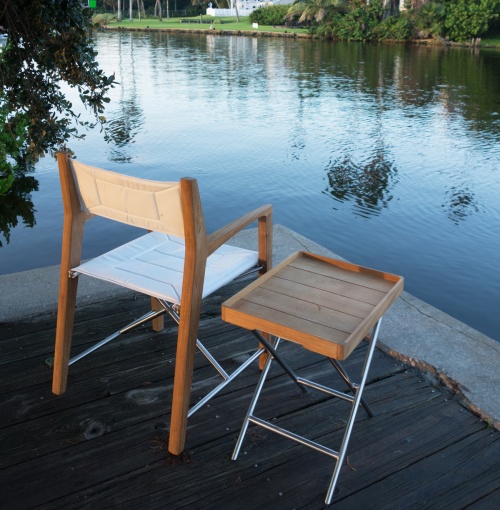 70461 odyssey teak and stainless steel folding chair and ottoman side table angled back view on dock with canal trees and building in background