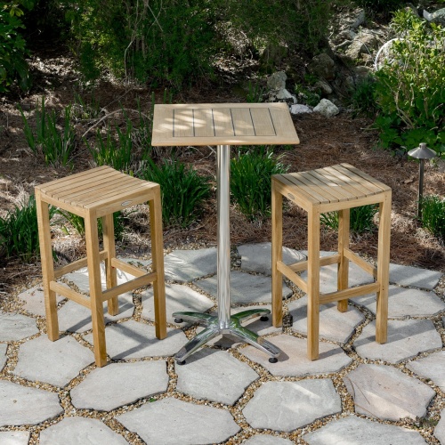 70670 Somerset teak Pub Bar Table and 2 teak Backless Barstools on stone patio with landscape shrubs and trees in background