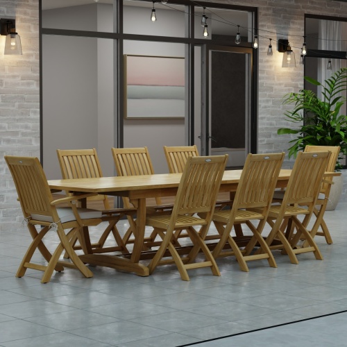 70794 Veranda Barbuda Teak 9 pc Dining Set of Grand Veranda Dining Table and 2 Barbuda Armchairs and 6 Side Chairs angled side view on a patio with patio lights and glass doors in background