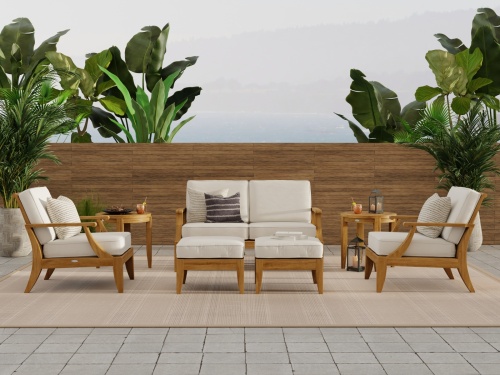 70877 Laguna 7 piece teak Love Seat Set on travertine tile patio open magazine on ottoman two pillar candles on side table potted palm tree with trees and house in  background