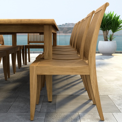 70917 Laguna 15 pc Teak Dining Set of Laguna 11 ft Rectangular Table and 14 Side Chairs in angled side view on tiled patio next to glass door with palm tree and ocean in background