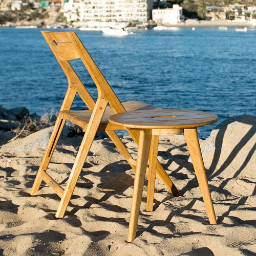 11916 Surf Teak Folding Side Chair and Side Table on sandy beach by boulders with water view and boats and buildings in back  