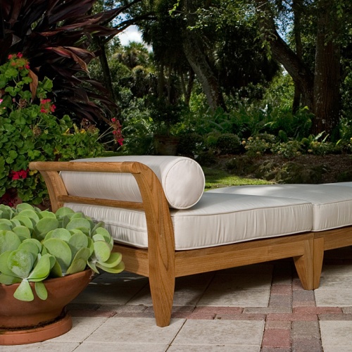 16766DP Aman Dais End Sectional and ottoman set angle view on a outside paver patio with a bolster and cushions with a container of green succulents in background of lush green shrubs