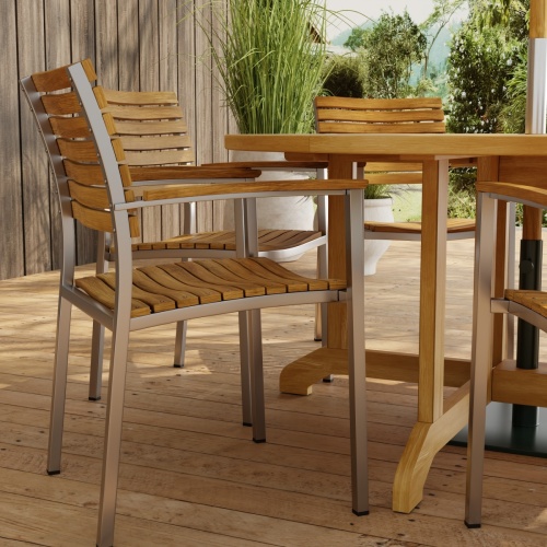 70349 Barbuda Vogue Teak 7 pc Dining Set showing closeup view of Vogue Teak and Stainless Steel  Armchairs in side view on paver patio with wood privacy wall and trees in background 
