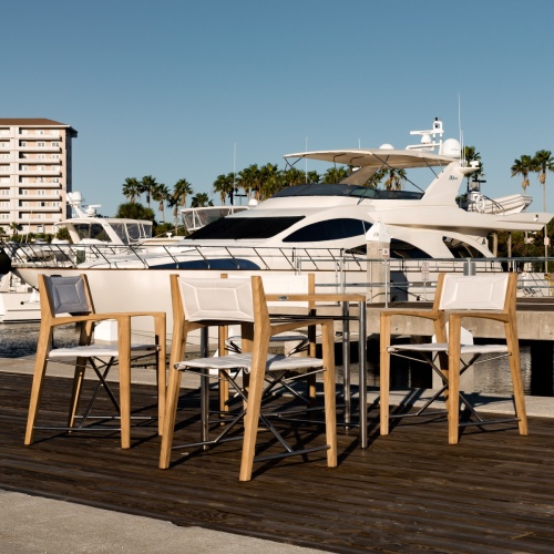70459 Odyssey 5 Piece folding teak and stainless steel dining set of Odyssey 32 inch square dining table and 4 Odyssey folding chairs on beach with ocean and sky in background