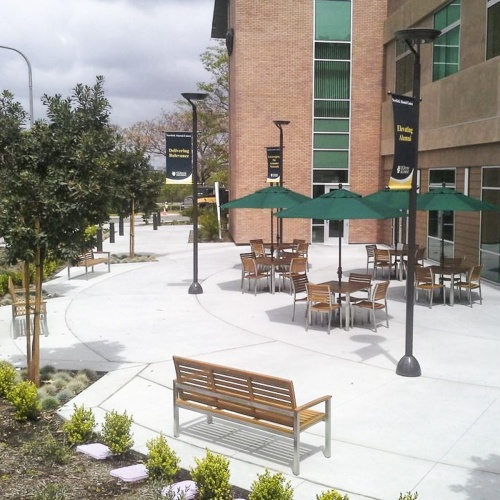 70443 Vogue 5 piece Bistro Square Dining Set showing 4 sets with optional opened market umbrellas on curved concrete patio next to a brick building surrounded by landscaping plants in background