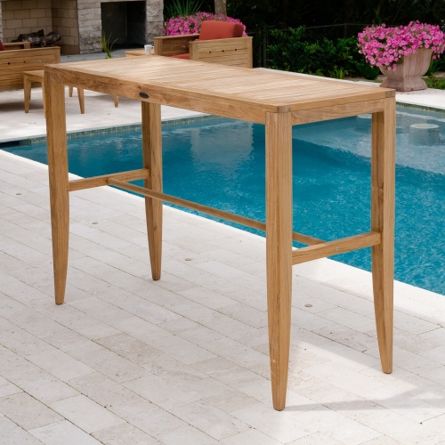 15821 Laguna Teak 5 ft  Bar Table on pavers next to pool with fireplace two craftsman chairs two potted flowering plants and shrubs in background