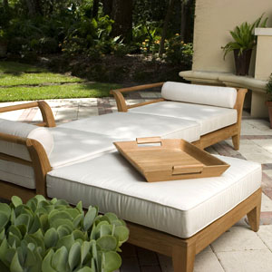 17440 folding teak tray table top angled view sitting on an aman dais sectional set surrounded by two potted plants with grass lawn and shrubs in background