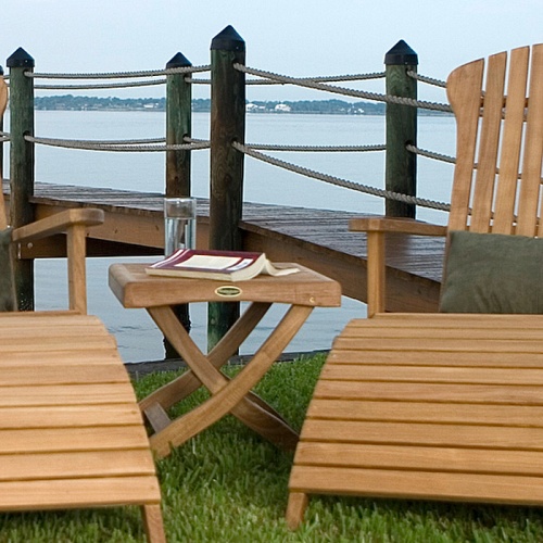 18600 Barbuda Ottoman Side Table with open book face down and glass of water on grass between two adirondack chairs with dock and lake in background