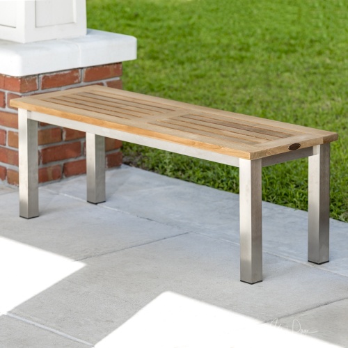 23940 Vogue Teak and Stainless Steel Backless Bench on concrete stone walkway next to brick and white pillar with grass lawn in background