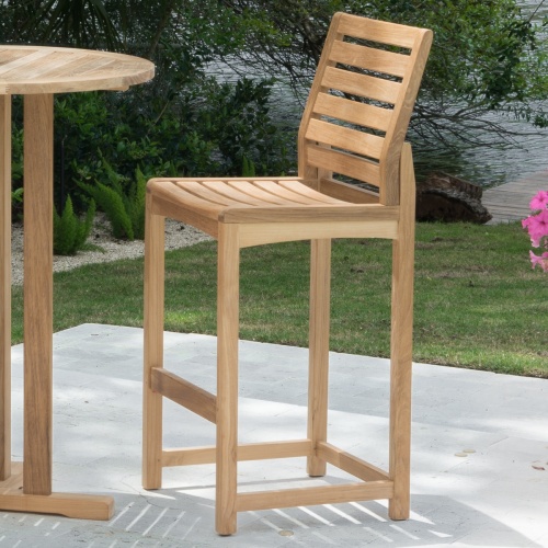 11812 Somerset Side Barstool and 30 inch Teak Bar Table on concrete patio with landscape plants lake and trees in background