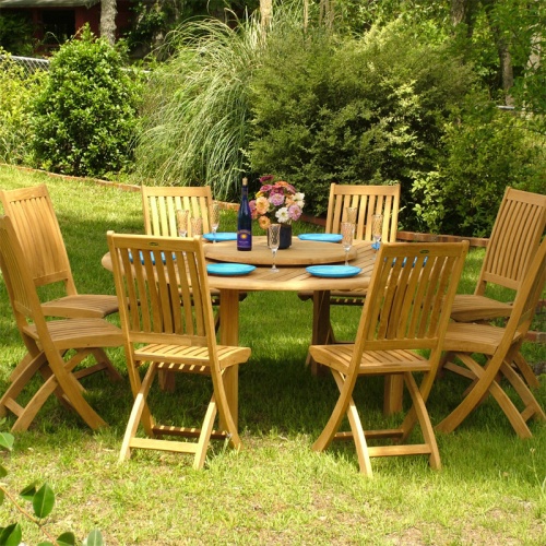 70018 Buckingham Barbuda 9 pc Dining Set angled with optional Lazy Susan with flower vase and blue plates and wine bottle and glasses on a grass with plants and fence in the back