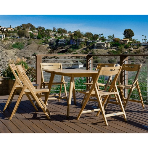 15917 Surf Teak Dining Table in angled end view on wood deck with railing overlooking homes along a hillside in background