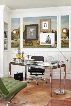 An image of a glass and metal office desk and chair showing framed photos and artwork in the background facing a green chair.